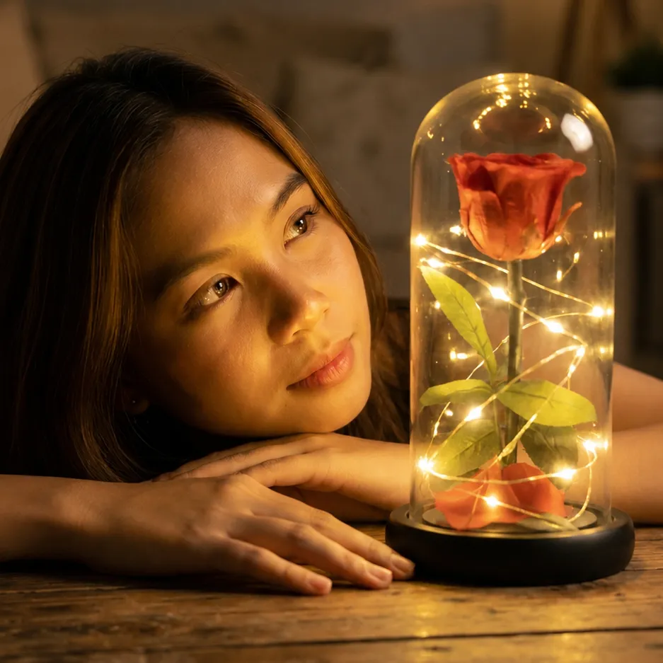 A young woman with dark hair rests her hands and head on a wooden table, gazing warmly at an illuminated glass dome beside her. Inside the clear cloche, a single vibrant red rose with green leaves is wrapped in delicate, glowing LED fairy lights on a dark base, casting a soft, golden light across her face in an intimate setting.
