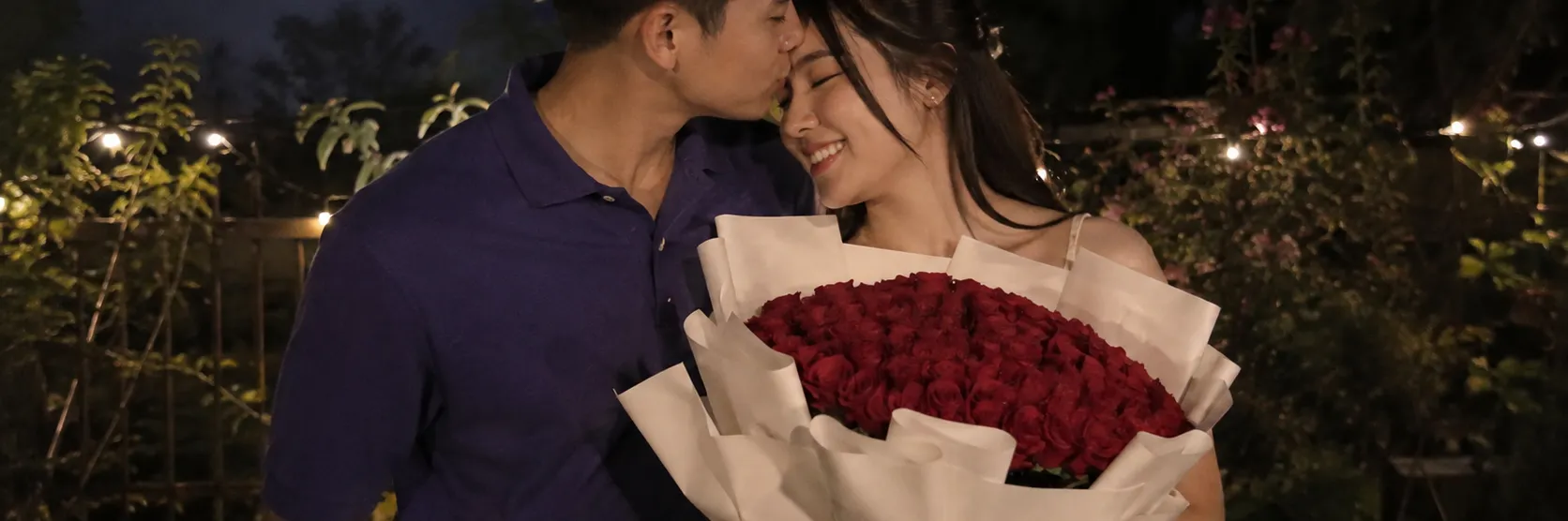 A young man in a blue polo shirt gently kisses a woman on her forehead outdoors at night. The woman smiles happily with her eyes closed while holding a large bouquet of vibrant red roses wrapped in white paper. Warm string lights glow softly among garden plants and dark foliage in the background.