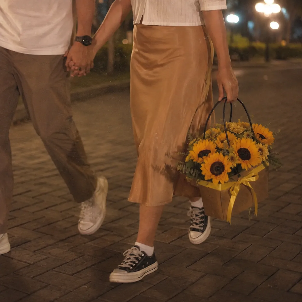 A couple walks hand-in-hand along a paved street at night, framed from the waist down. The woman wears a satin gold midi skirt with black canvas sneakers and carries a brown gift basket filled with bright yellow sunflowers tied with a yellow ribbon. Beside her, the man wears khaki pants and white sneakers under warm evening streetlights.