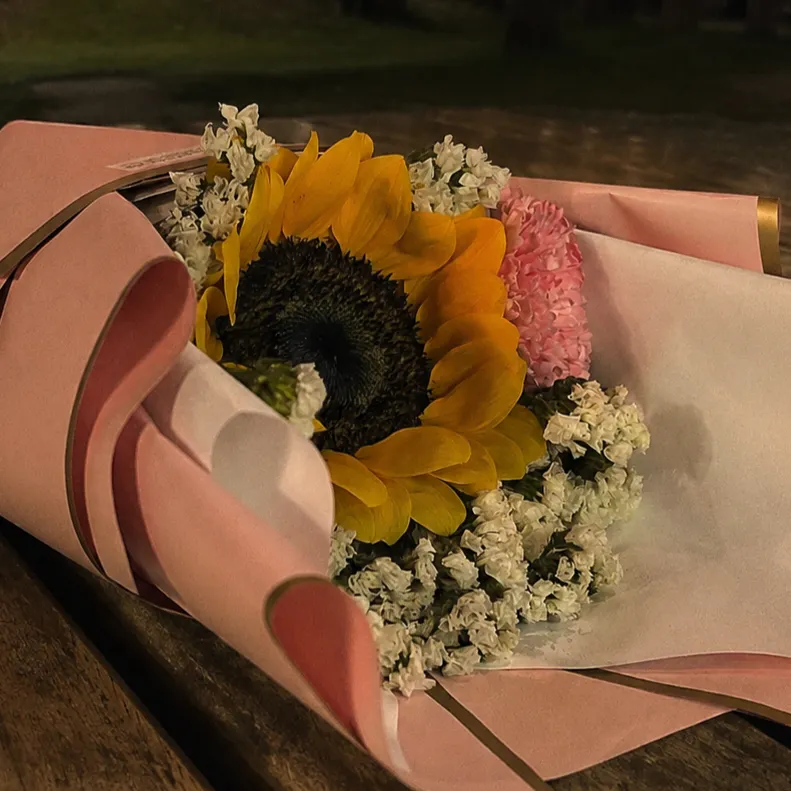 A floral bouquet rests horizontally on a rustic wooden surface in soft evening lighting. The arrangement features a prominent yellow sunflower surrounded by small white filler flowers and pink blossoms, neatly wrapped in light pink paper with dark-trimmed borders.