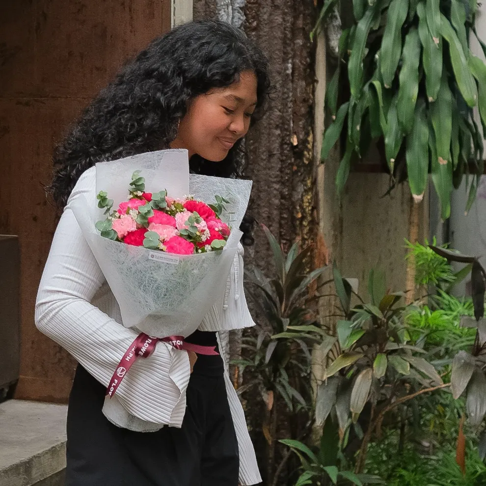 A young man stands with his arm wrapped around a woman as they both look down at a bouquet of pink, white, and green carnations wrapped in light pink paper. He wears a black jacket and dark shirt, while she wears a purple floral-patterned blouse and smiles softly at the arrangement in her hands.