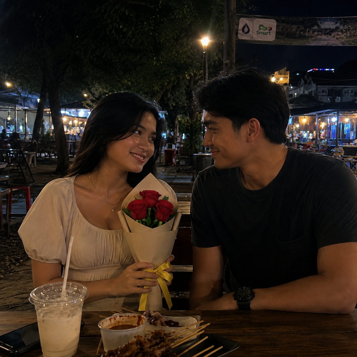 A young couple sits together at an outdoor table at night, gazing warmly at each other. The woman wears a white puff-sleeve top and holds a small bouquet of red roses tied with a yellow ribbon. The man, wearing a dark t-shirt, rests his arm on the wooden table, which holds grilled food skewers, dipping sauce, and a cold drink in a clear plastic cup under soft ambient streetlights.