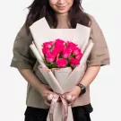 Woman holding bouquet of 12 vibrant pink roses wrapped in white paper with pink ribbon