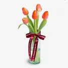 Five red-orange tulips with green stems in a clear vase with burgundy ribbon