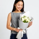 White roses bouquet with green foliage wrapped in gray paper and white ribbon