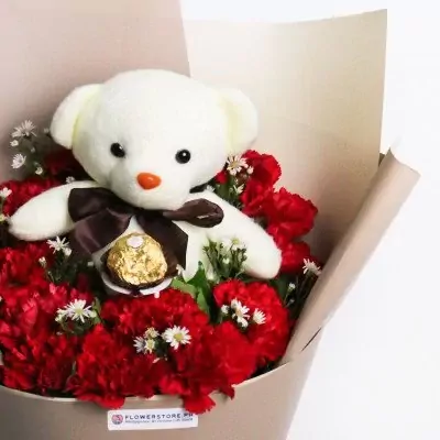 White teddy bear with black bow tie surrounded by red carnations and white baby's breath flowers