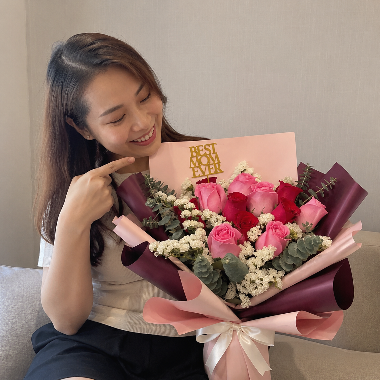 A smiling young woman with long brown hair sits on a couch, looking down and pointing at a large, vibrant bouquet of flowers she is holding. The bouquet features a mix of pink and deep red roses, small white filler flowers, and green eucalyptus leaves, all beautifully wrapped in layers of dark purple and light pink paper tied with a white satin ribbon. Poked into the back of the arrangement is a square pink card topped with a decorative gold cutout that reads Best Mom Ever.