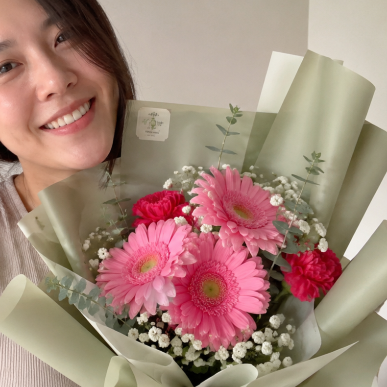 A close-up shot shows a young woman smiling brightly alongside a large, beautifully arranged floral bouquet. The bouquet features three prominent pink gerbera daisies in the center, accented by deep red carnations, delicate white baby's breath, and sprigs of eucalyptus foliage. The flowers are wrapped in layers of sage green paper, with a small square sticker visible on the wrapping.