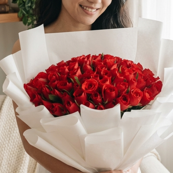 A smiling woman holds a massive, rounded bouquet of deep red roses, which are strikingly wrapped in multiple layers of crisp, flared white paper. The stark contrast between the vibrant red blooms and the clean white wrapping creates a bold, modern, and elegant presentation.