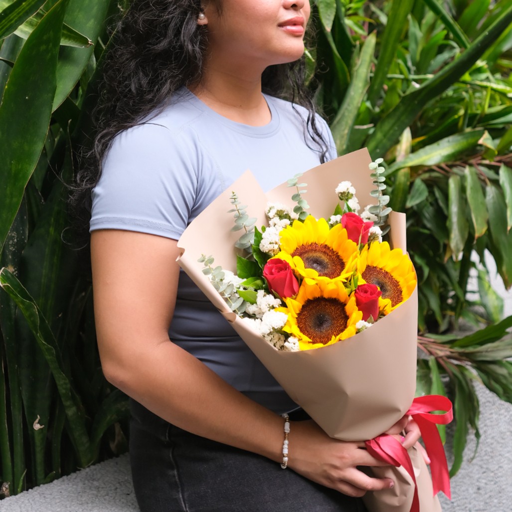 A woman in a blue top holds a cheerful bouquet featuring three large sunflowers and vibrant red roses. The arrangement is accented with white baby's breath and silver-green eucalyptus sprigs, all neatly wrapped in matte tan paper and tied with a bright red satin bow. The candid lifestyle shot is set against a lush backdrop of green tropical plants, creating a natural and warm aesthetic.