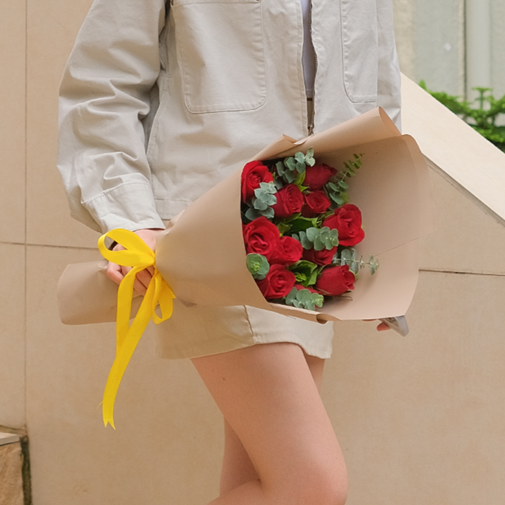 A person wearing a light beige jacket and a matching short skirt stands outside, holding a bouquet of vibrant red roses. The roses are interspersed with sprigs of green eucalyptus, wrapped in smooth light brown kraft paper, and securely tied with a bright yellow satin ribbon that drapes downward. The background features a textured beige stone wall and a hint of green foliage.