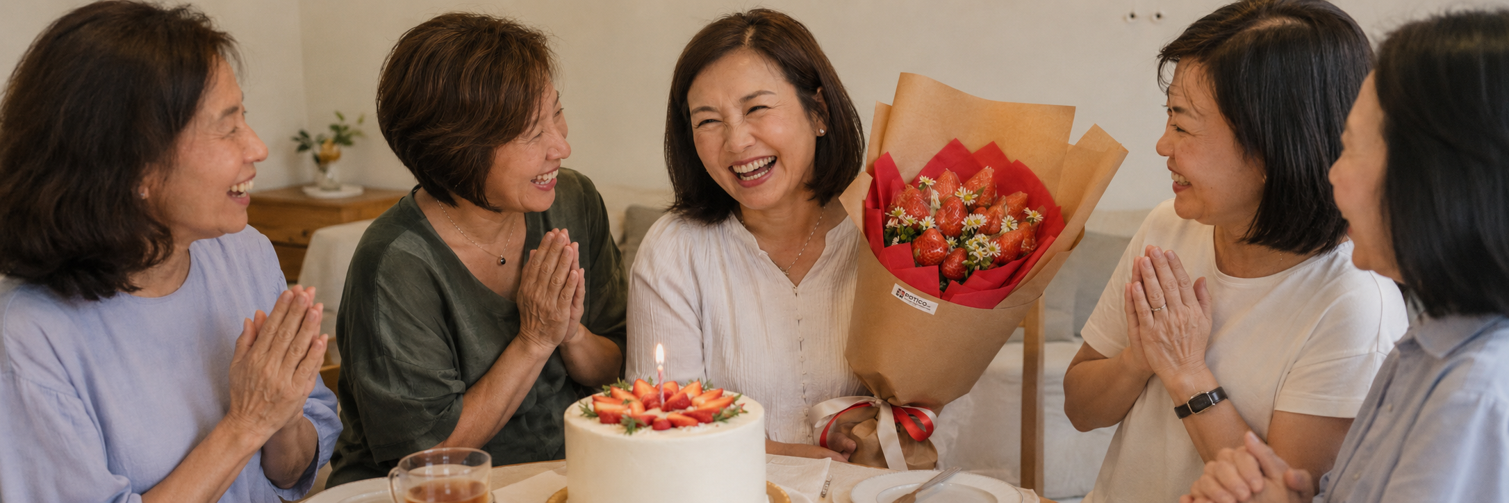 A group of five middle-aged Singaporean women are gathered around a table celebrating a birthday in a brightly lit, cozy indoor setting. The woman in the center laughs joyfully, holding a large bouquet made of fresh strawberries and small white chamomile flowers wrapped in brown craft paper with a red ribbon. In front of her sits a round white frosted cake topped with sliced strawberries and a single lit candle. The other four women, two on her left and two on her right, look toward her with warm, happy expressions, their hands pressed together in appreciative gestures. They are dressed in casual, comfortable blouses in shades of blue, olive green, white, and grey, creating a warm, intimate atmosphere of friendship and celebration.