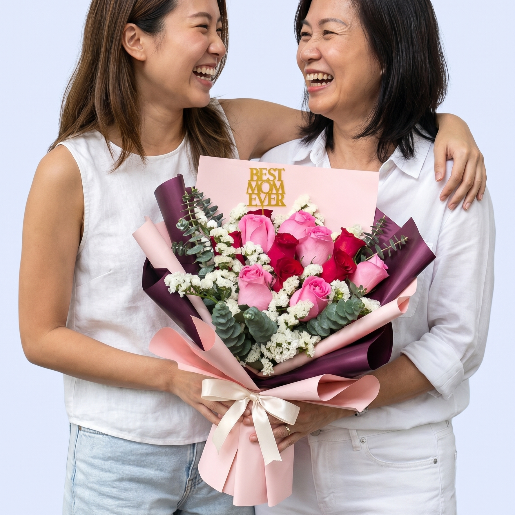 A candid studio shot captures a joyful moment between a younger and an older woman, both dressed in white sleeveless tops and laughing together. They are holding a large, elegant bouquet of pink and red roses accented with white baby's breath and eucalyptus sprigs. The arrangement is wrapped in layered light pink and deep purple paper, tied with a cream satin bow, and features a gold
