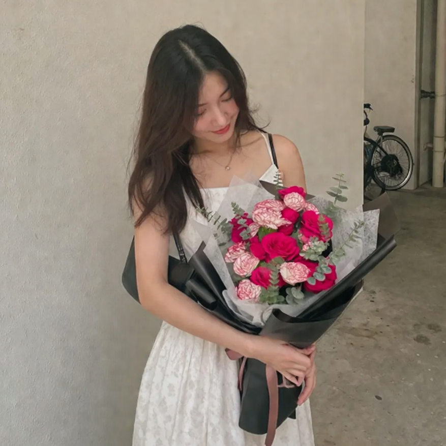 A woman in a white patterned dress holds a sophisticated bouquet of bright red roses and variegated pink-and-white carnations. The arrangement is accented with silver-green eucalyptus sprigs and wrapped in sleek, flared black paper, creating a modern and high-contrast floral gift.