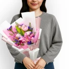 A woman in a grey sweater holds a bouquet of pink lilies, roses, and purple statice.
