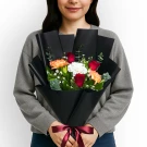 A person holds a bouquet of red roses, orange and white carnations, and greenery wrapped in black paper, tied with a red ribbon.