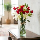 Red roses and white lilies in a clear glass vase with twine tie, on a wooden table.