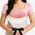 Woman in pink top holding a large bouquet of pink carnations wrapped in white paper, tied with a black ribbon.