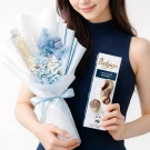 Woman holds a blue dried flower bouquet and a box of Belgian chocolate seashells.
