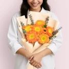 Woman holding a bouquet of bright orange gerbera daisies wrapped in peach paper.