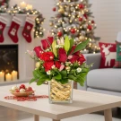 Red roses, carnations, and lilies in a glass vase on a table, with a Christmas tree and fireplace in the background.