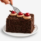 Slice of chocolate cake with cherries and whipped cream being cut by a fork.