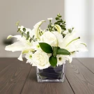 White roses, lilies, and daisies in a clear vase with greenery on a wooden table.