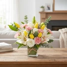 A bouquet of yellow tulips, white lilies, and pink carnations in a square vase on a wooden table.