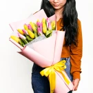 A young woman in a rust-colored top holds a large bouquet of pink and yellow tulips, tied with a yellow ribbon.
