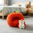 A plush hedgehog toy sits beside an orange apple-shaped pet bed on a carpeted floor, with a bed in the background.