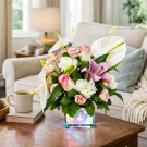 Pink and white floral arrangement in a square glass vase with fairy lights, beside a mug of coffee and a book.