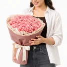 A person holding a bouquet of 60 pink carnations wrapped in blush paper with a white ribbon.