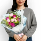 A person in a grey sweater holds a bouquet of pink roses, green button mums, and golden phoenix flowers.