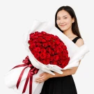A woman smiles holding a large bouquet of 99 red roses wrapped in white paper and tied with a red ribbon.