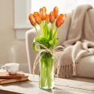 Red-orange tulips in a clear glass vase tied with twine, with a steaming cup of tea and books in the background.