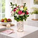 A vase of flowers with fairy lights, a candle, and a fruit bowl on a table.