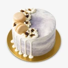 A marbelized purple and white cake with white drip frosting, macarons, and floral decorations on a gold cake board.