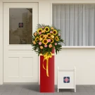 Sunburst Celebration arrangement with sunflowers and pink/orange carnations in a red stand.