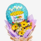 A birthday balloon with a cake design sits atop a bouquet of yellow gerbera daisies and small white flowers.