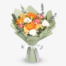 A bright bouquet of orange gerberas, carnations, and white flowers wrapped in green paper with a white ribbon.