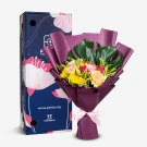 A flower bouquet with yellow, pink, and fuchsia blooms, wrapped in purple paper, next to a blue gift box.