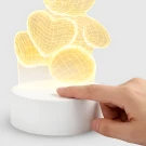 A finger presses the power button on a white base below a glowing teddy bear lamp with heart shapes.