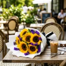 Bouquet of sunflowers and purple statice flowers wrapped in white paper, tied with a black ribbon.