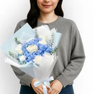 A person holds a bouquet of blue hydrangeas, white roses, and carnations, wrapped in blue and white paper.