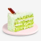 Slice of green ondeh ondeh cake with white frosting and shredded coconut, on a pink plate with a dark brown syrup topping.