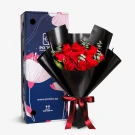 A bouquet of 12 red roses in black paper with a burgundy ribbon, next to a dark blue gift box with pink floral designs.