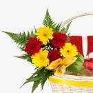Basket of red and yellow gerbera daisies, red carnations, ferns, fruit, and a red knitted item.