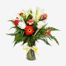 A vibrant bouquet of white lilies, orange gerberas, red roses, and greenery in a clear glass vase with a yellow ribbon.