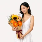 Woman in white shirt holds a bouquet with a sunflower, orange gerberas, and yellow carnations.