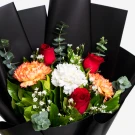 A vibrant bouquet of red roses, orange carnations, and white blooms with green foliage, wrapped in black paper.